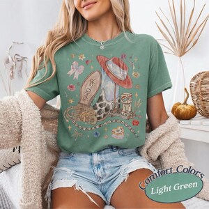 May include: A light green short-sleeve t-shirt featuring a graphic print of a cowboy boot, a red hat, a belt, and blocks with the number 13. The boot has the name Taylor printed on it. The design includes small floral accents and stars.