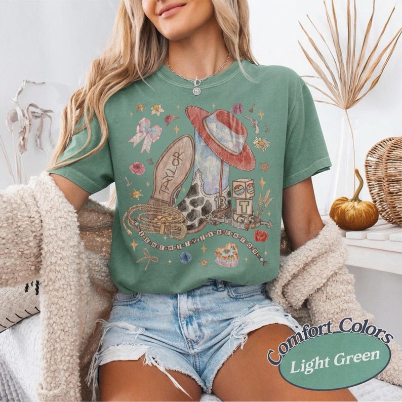 May include: A light green short-sleeve t-shirt featuring a graphic print of a cowboy boot, a red hat, a belt, and blocks with the number 13. The boot has the name Taylor printed on it. The design includes small floral accents and stars.