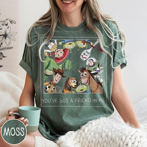 May include: Green t-shirt featuring a colorful graphic of characters from the animated film Toy Story. The graphic includes Woody, Buzz Lightyear, Rex, and other characters. The text "YOU'VE GOT A FRIEND IN ME" is printed below the graphic.