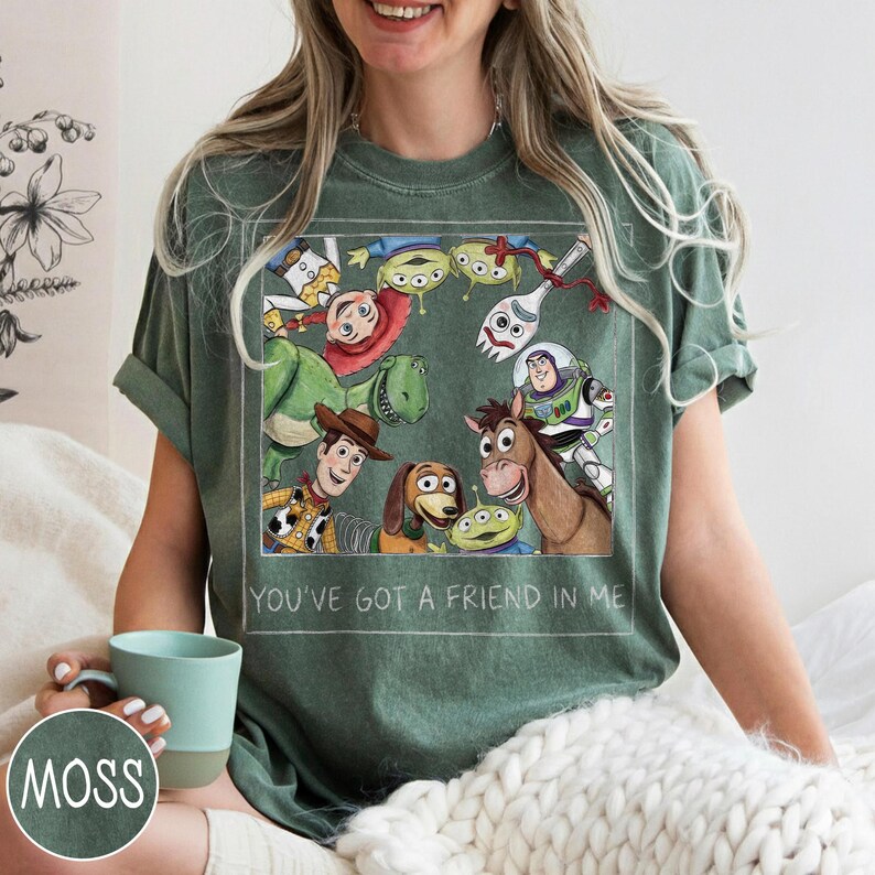 May include: Green t-shirt featuring a colorful graphic of characters from the animated film Toy Story. The graphic includes Woody, Buzz Lightyear, Rex, and other characters. The text "YOU'VE GOT A FRIEND IN ME" is printed below the graphic.