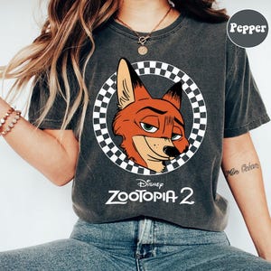 May include: Dark gray t-shirt featuring a cartoon image of Nick Wilde from Disney's Zootopia 2. The fox's head is inside a checkered circle. The words "Disney Zootopia 2" are printed below the image in white.