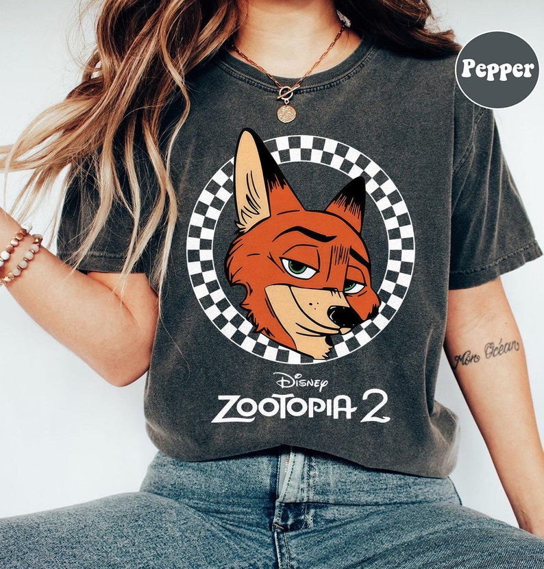 May include: Dark gray t-shirt featuring a cartoon image of Nick Wilde from Disney's Zootopia 2. The fox's head is inside a checkered circle. The words "Disney Zootopia 2" are printed below the image in white.