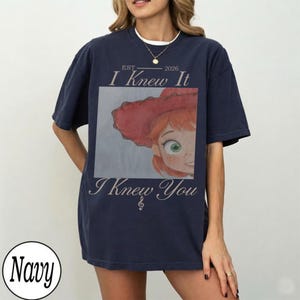 May include: A navy blue short-sleeve t-shirt featuring a graphic print of a character with red hair and a red hat. The text reads EST 2026 I Knew It I Knew You with a small musical note symbol. The design is centered on the front of the shirt.
