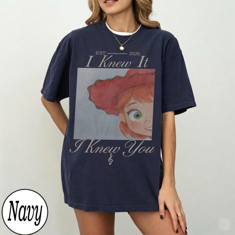 May include: A navy blue short-sleeve t-shirt featuring a graphic print of a character with red hair and a red hat. The text reads EST 2026 I Knew It I Knew You with a small musical note symbol. The design is centered on the front of the shirt.