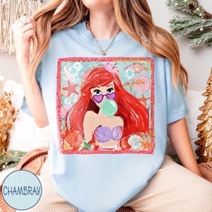 May include: Light blue t-shirt featuring a colorful graphic of a cartoon mermaid with red hair, heart-shaped sunglasses, and a bubblegum bubble. The design includes seashells, starfish, and fish. The shirt has the word "Chambray" on the bottom left.