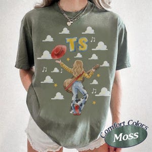 May include: An olive green t-shirt featuring a graphic of a person with long hair playing an acoustic guitar. The design includes a red hat, white clouds, yellow stars, musical notes, and the yellow text TS. The person wears a plaid shirt and cow print pants.