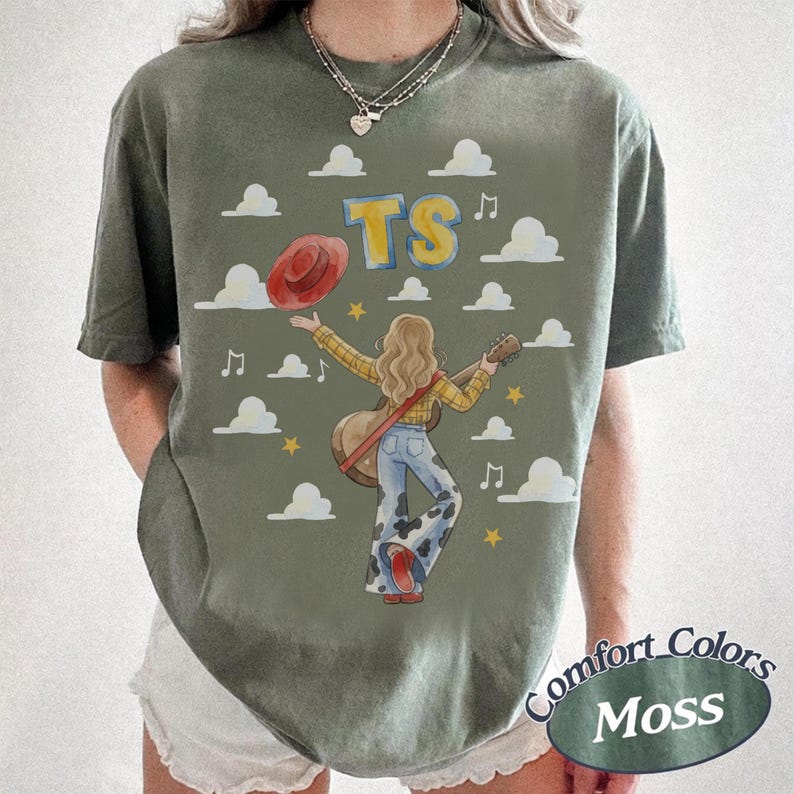 May include: An olive green t-shirt featuring a graphic of a person with long hair playing an acoustic guitar. The design includes a red hat, white clouds, yellow stars, musical notes, and the yellow text TS. The person wears a plaid shirt and cow print pants.