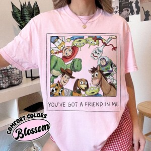 May include: Pink t-shirt featuring a graphic of Toy Story characters within a black border. The shirt includes the text "YOU'VE GOT A FRIEND IN ME" and the Comfort Colors Blossom logo.