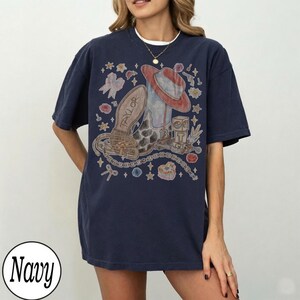 May include: A navy blue short-sleeve t-shirt featuring a graphic print of a cowboy boot, a hat, a belt, and a block with the letter T. The design includes small stars, flowers, and bows surrounding the central items, with the name Taylor on the boot.