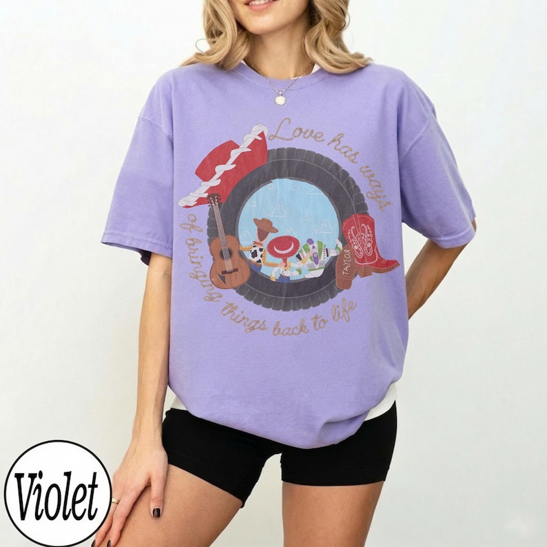 K&ouml;nnte beinhalten: Ein hellviolettes Kurzarm-T-Shirt mit einer Grafik eines Reifens, der eine Gitarre, einen roten Cowboyhut und einen roten Cowboystiefel umrahmt. Der Text "Love has ways of bringing things back to life" ist in Schreibschrift aufgedruckt.