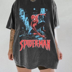 May include: Dark gray oversized t-shirt featuring a graphic of Spider-Man in a dynamic pose against a cityscape backdrop. The word "SPIDERMAN" is printed in red, stylized lettering. The shirt has short sleeves and a relaxed fit.