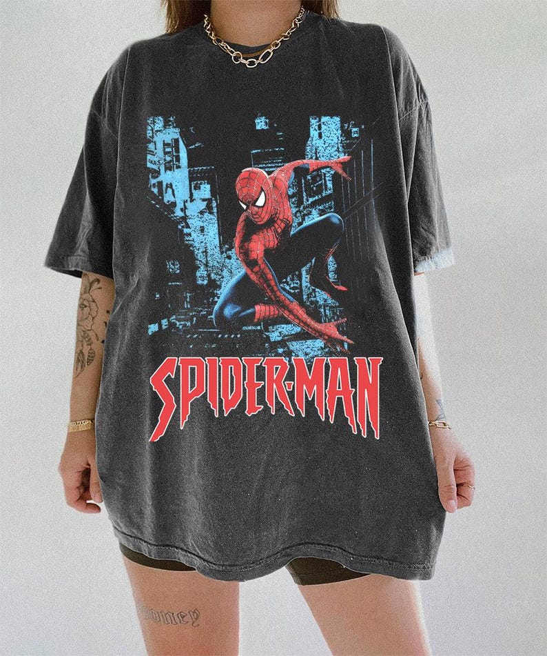 May include: Dark gray oversized t-shirt featuring a graphic of Spider-Man in a dynamic pose against a cityscape backdrop. The word "SPIDERMAN" is printed in red, stylized lettering. The shirt has short sleeves and a relaxed fit.