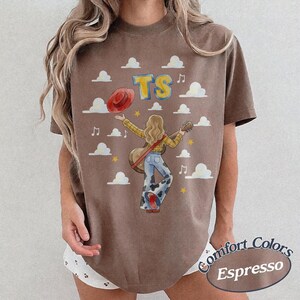 May include: A brown short-sleeve t-shirt featuring a graphic of a person with long hair holding a guitar and a red hat. The design includes the letters TS, white clouds, musical notes, and yellow stars. The shirt has a crew neckline and a relaxed fit.