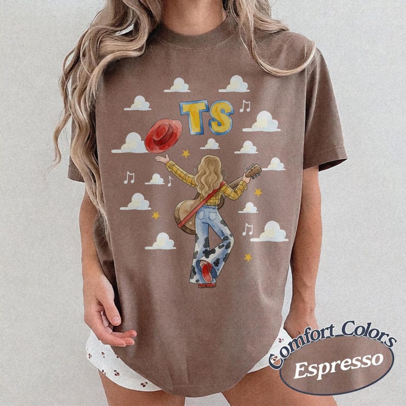May include: A brown short-sleeve t-shirt featuring a graphic of a person with long hair holding a guitar and a red hat. The design includes the letters TS, white clouds, musical notes, and yellow stars. The shirt has a crew neckline and a relaxed fit.