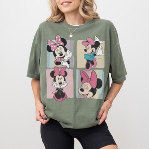 Op de afbeelding: Olijfgroen T-shirt met vier panelen met Minnie Mouse-illustraties. Elk paneel toont een andere pose van Minnie, met haar kenmerkende strik en verschillende outfits. Het T-shirt heeft een relaxte pasvorm, perfect voor casual wear.