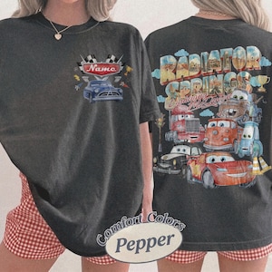 May include: A charcoal gray t-shirt featuring a small graphic on the front chest with a blue car and the text Name. The back displays a large, colorful graphic of various cartoon cars with the text Radiator Springs and Lightning McQueen.