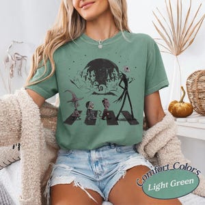 May include: A light green short-sleeve t-shirt featuring a black graphic print of characters from The Nightmare Before Christmas walking across a crosswalk in front of a large moon and a spooky town silhouette, with a crew neckline.
