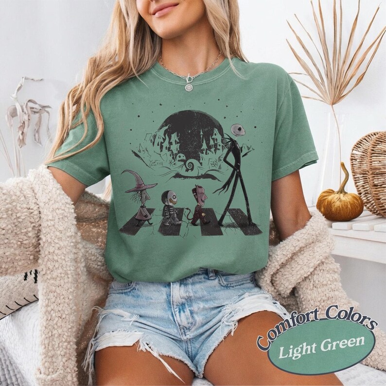 May include: A light green short-sleeve t-shirt featuring a black graphic print of characters from The Nightmare Before Christmas walking across a crosswalk in front of a large moon and a spooky town silhouette, with a crew neckline.
