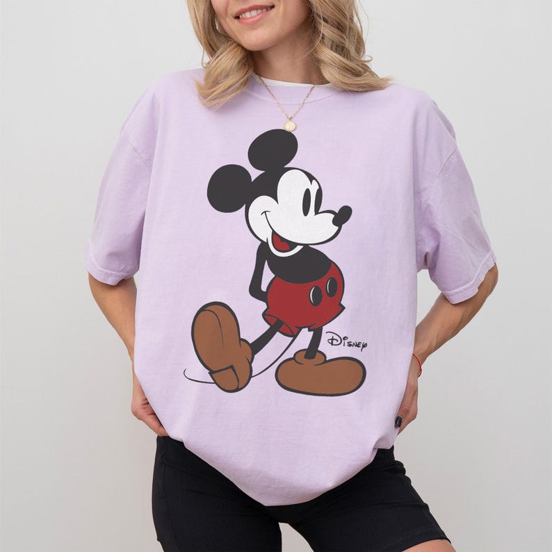 May include: A light purple t-shirt featuring a classic Mickey Mouse graphic. The cartoon character is depicted in black, red, and brown, with the Disney logo printed below. The shirt has short sleeves and a relaxed fit.