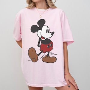 May include: A light pink t-shirt featuring a large graphic of Mickey Mouse. The cartoon character is depicted in his classic outfit: black head and ears, white face, red shorts, and brown shoes. The Disney logo is printed on the shirt.