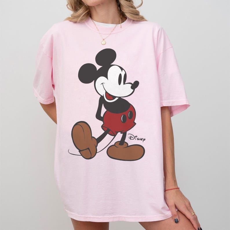 May include: A light pink t-shirt featuring a large graphic of Mickey Mouse. The cartoon character is depicted in his classic outfit: black head and ears, white face, red shorts, and brown shoes. The Disney logo is printed on the shirt.