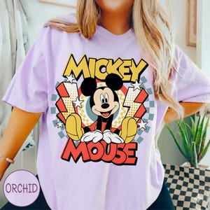 May include: A light purple t-shirt featuring a graphic of Mickey Mouse. The design includes the words "MICKEY" and "MOUSE" in yellow and red, with Mickey in the center. The background has stars, lightning bolts, and a checkered pattern.
