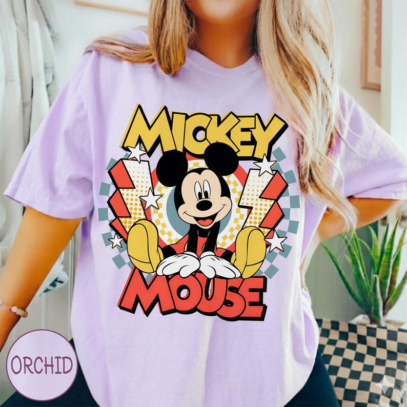 May include: A light purple t-shirt featuring a graphic of Mickey Mouse. The design includes the words "MICKEY" and "MOUSE" in yellow and red, with Mickey in the center. The background has stars, lightning bolts, and a checkered pattern.