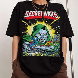 Puede incluir: Camiseta negra con una ilustraci&oacute;n de c&oacute;mic. El texto "SECRET WARS" est&aacute; en blanco y rojo. El gr&aacute;fico muestra un personaje con rostro met&aacute;lico y capucha verde, sosteniendo varios personajes de c&oacute;mic en su mano.