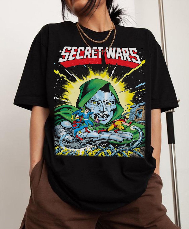Puede incluir: Camiseta negra con una ilustraci&oacute;n de c&oacute;mic. El texto "SECRET WARS" est&aacute; en blanco y rojo. El gr&aacute;fico muestra un personaje con rostro met&aacute;lico y capucha verde, sosteniendo varios personajes de c&oacute;mic en su mano.