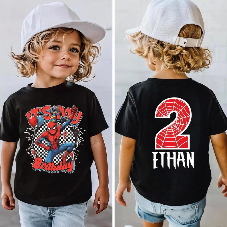 May include: Black t-shirt featuring a Spider-Man graphic with the text "It's My Birthday" and a checkered background. The back of the shirt displays a red number 2 with a spiderweb design and the name "Ethan" in white. The child is wearing a white baseball cap.
