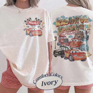 May include: An ivory t-shirt featuring a small graphic on the front with a red race car and the text Name, and a large back graphic displaying multiple animated car characters, the text Radiator Springs and Lightning McQueen, and clouds.