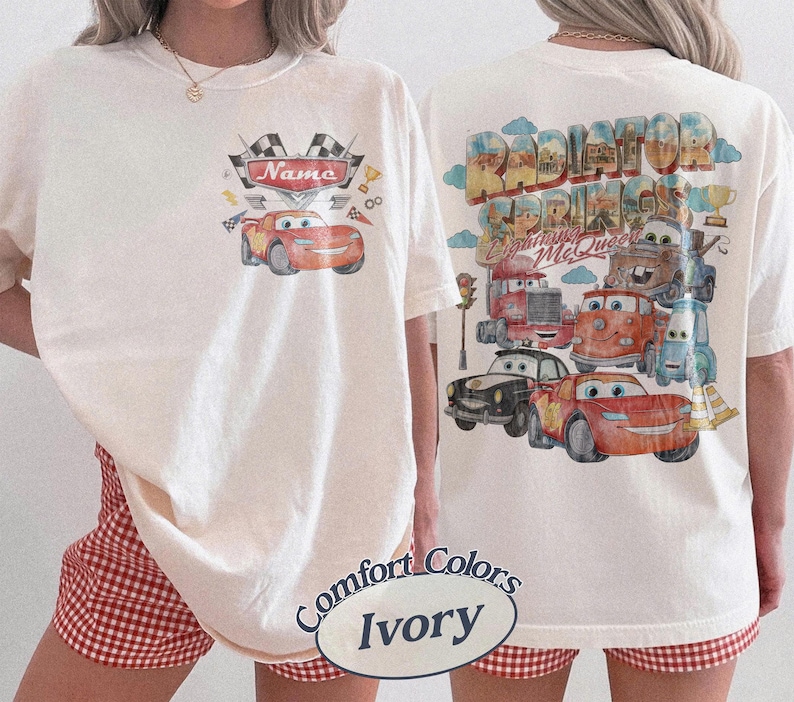 May include: An ivory t-shirt featuring a small graphic on the front with a red race car and the text Name, and a large back graphic displaying multiple animated car characters, the text Radiator Springs and Lightning McQueen, and clouds.