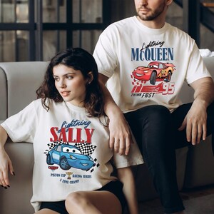May include: Two off-white t-shirts with graphics from the film "Cars." One shows "Lightning McQueen" with a red race car, the other "Lightning Sally" with a blue race car. Both shirts include text and racing-themed designs, perfect for fans of the animated movie.