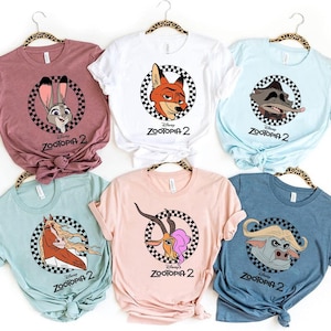 May include: Assorted-color t-shirts featuring characters from Zootopia 2. Each shirt displays a different character within a black and white checkered circle, with the movie title below. The shirts are in various colors, including pink, white, and blue.