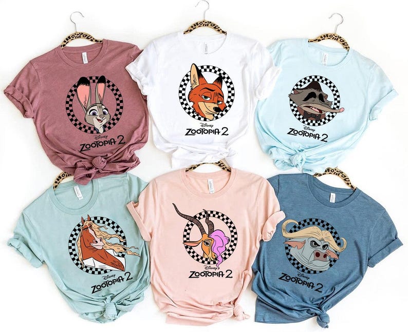 May include: Assorted-color t-shirts featuring characters from Zootopia 2. Each shirt displays a different character within a black and white checkered circle, with the movie title below. The shirts are in various colors, including pink, white, and blue.
