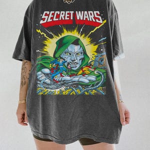 Puede incluir: Camiseta gris oscuro con una ilustraci&oacute;n de c&oacute;mic y el texto "SECRET WARS". El gr&aacute;fico muestra una escena de batalla con personajes y el Doctor Doom. La camiseta tiene mangas cortas y un corte holgado.