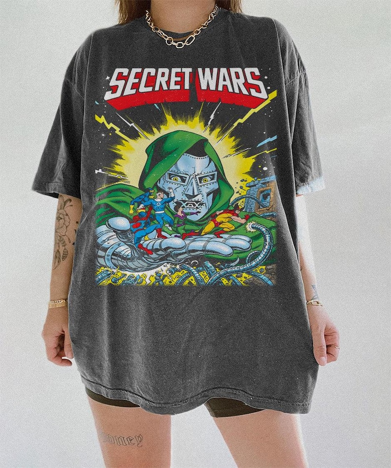 Puede incluir: Camiseta gris oscuro con una ilustraci&oacute;n de c&oacute;mic y el texto "SECRET WARS". El gr&aacute;fico muestra una escena de batalla con personajes y el Doctor Doom. La camiseta tiene mangas cortas y un corte holgado.