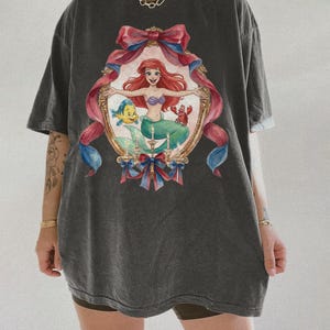 May include: Dark gray t-shirt featuring a framed illustration of a mermaid with red hair, a purple seashell top, and a green tail. The frame is decorated with pink and blue ribbons. The shirt is oversized.
