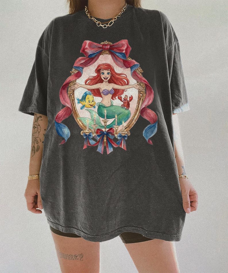 May include: Dark gray t-shirt featuring a framed illustration of a mermaid with red hair, a purple seashell top, and a green tail. The frame is decorated with pink and blue ribbons. The shirt is oversized.