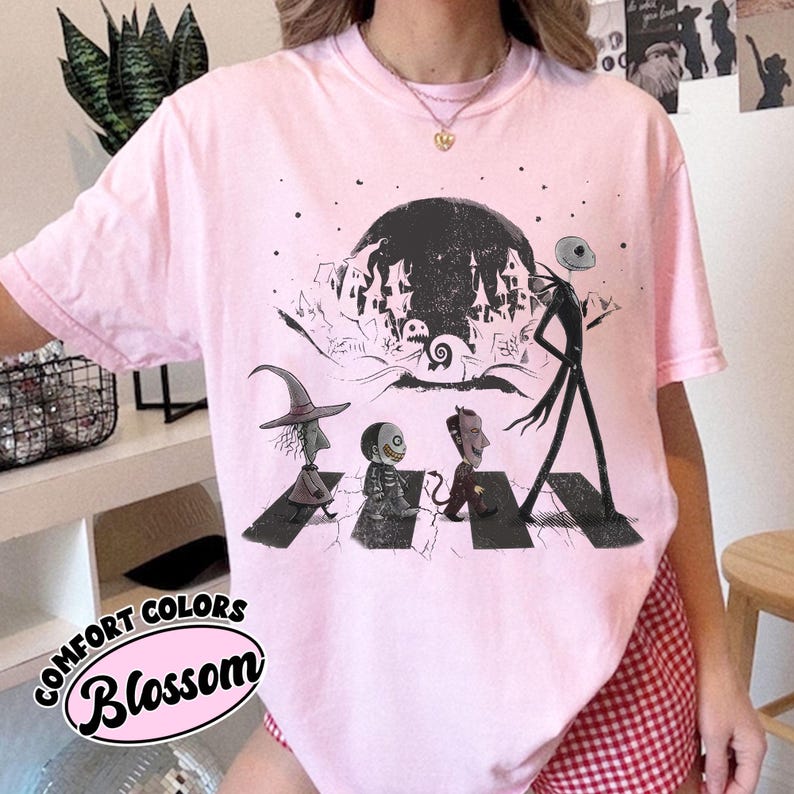 May include: A light pink t-shirt featuring a graphic print of Jack Skellington and three trick-or-treaters walking across a crosswalk. The design includes a large dark moon and a spooky town silhouette in the background, rendered in black ink.