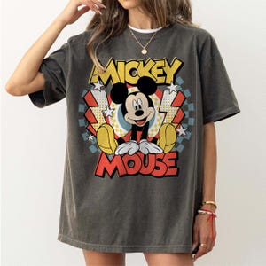 May include: Dark gray t-shirt featuring a colorful graphic of Mickey Mouse. The design includes the words "Mickey Mouse" in yellow and red, with a retro style. The shirt has short sleeves and a relaxed fit.