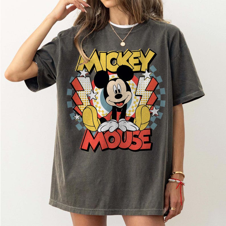 May include: Dark gray t-shirt featuring a colorful graphic of Mickey Mouse. The design includes the words "Mickey Mouse" in yellow and red, with a retro style. The shirt has short sleeves and a relaxed fit.