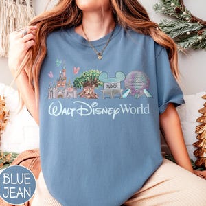May include: Blue jean coloured t-shirt featuring a Walt Disney World graphic. The graphic includes a castle, tree, and other park icons. The words "Walt Disney World" are written in a script font.