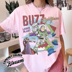 May include: A pale pink t-shirt with a graphic of Buzz Lightyear, the text "BUZZ Lightyear" and "LASER BLAST". The design includes Zurg and the Aliens from Toy Story. The shirt is from the Comfort Colors collection, with the colour "Blossom".