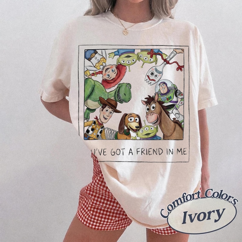 May include: Ivory-colored t-shirt featuring a rectangular graphic of Toy Story characters, including Woody, Buzz Lightyear, and Rex. The text "I'VE GOT A FRIEND IN ME" is below the graphic. The shirt is a Comfort Colors brand.