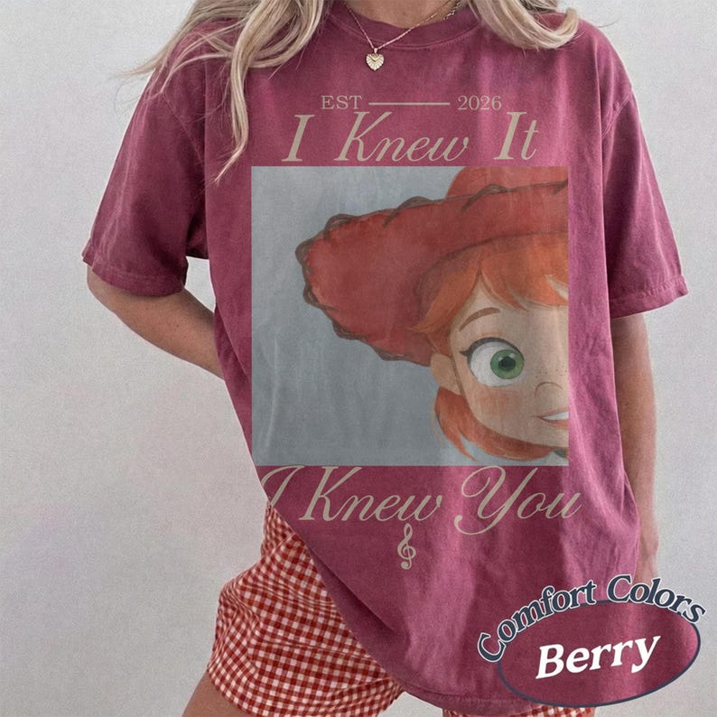 May include: A berry-colored short-sleeve t-shirt featuring a graphic of a cartoon character with red hair and a red hat. The text reads EST 2026 I Knew It I Knew You with a musical note symbol. The shirt has a vintage-style washed appearance.
