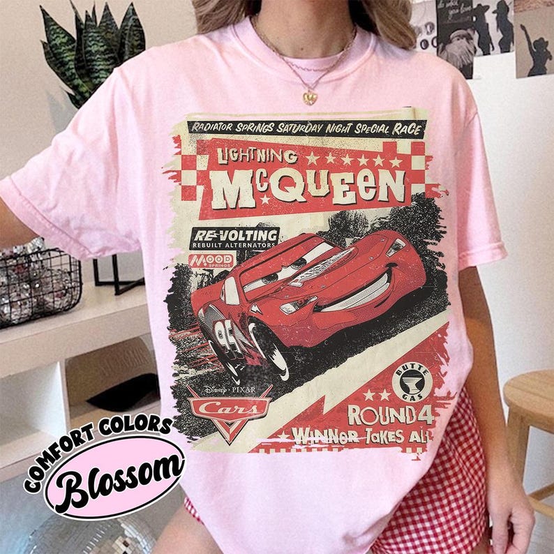 May include: A light pink t-shirt featuring a graphic of Lightning McQueen from the Disney Pixar movie Cars. The design includes the text "Lightning McQueen," "Radiator Springs," and "Round 4." The shirt is labeled "Comfort Colors Blossom."
