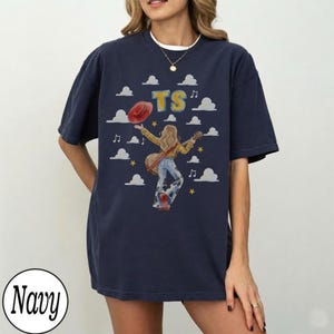 May include: A navy blue t-shirt featuring a graphic of a person with long hair wearing a yellow shirt and cow-print bell-bottom pants, holding a guitar. The design includes a red hat, yellow stars, white clouds, musical notes, and the letters TS.
