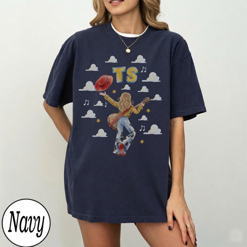 May include: A navy blue t-shirt featuring a graphic of a person with long hair wearing a yellow shirt and cow-print bell-bottom pants, holding a guitar. The design includes a red hat, yellow stars, white clouds, musical notes, and the letters TS.