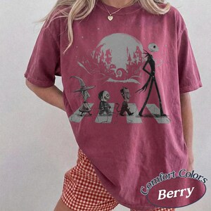 May include: A berry-colored short-sleeve t-shirt featuring a graphic print of four characters walking across a crosswalk in front of a large moon and a spooky castle silhouette. The design is rendered in shades of black, grey, and white.